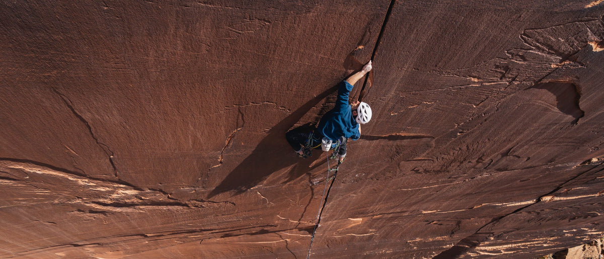 A high-angle action shot of a rock climber scaling a massive red sandstone cliff face, wearing a blue jacket and white helmet.