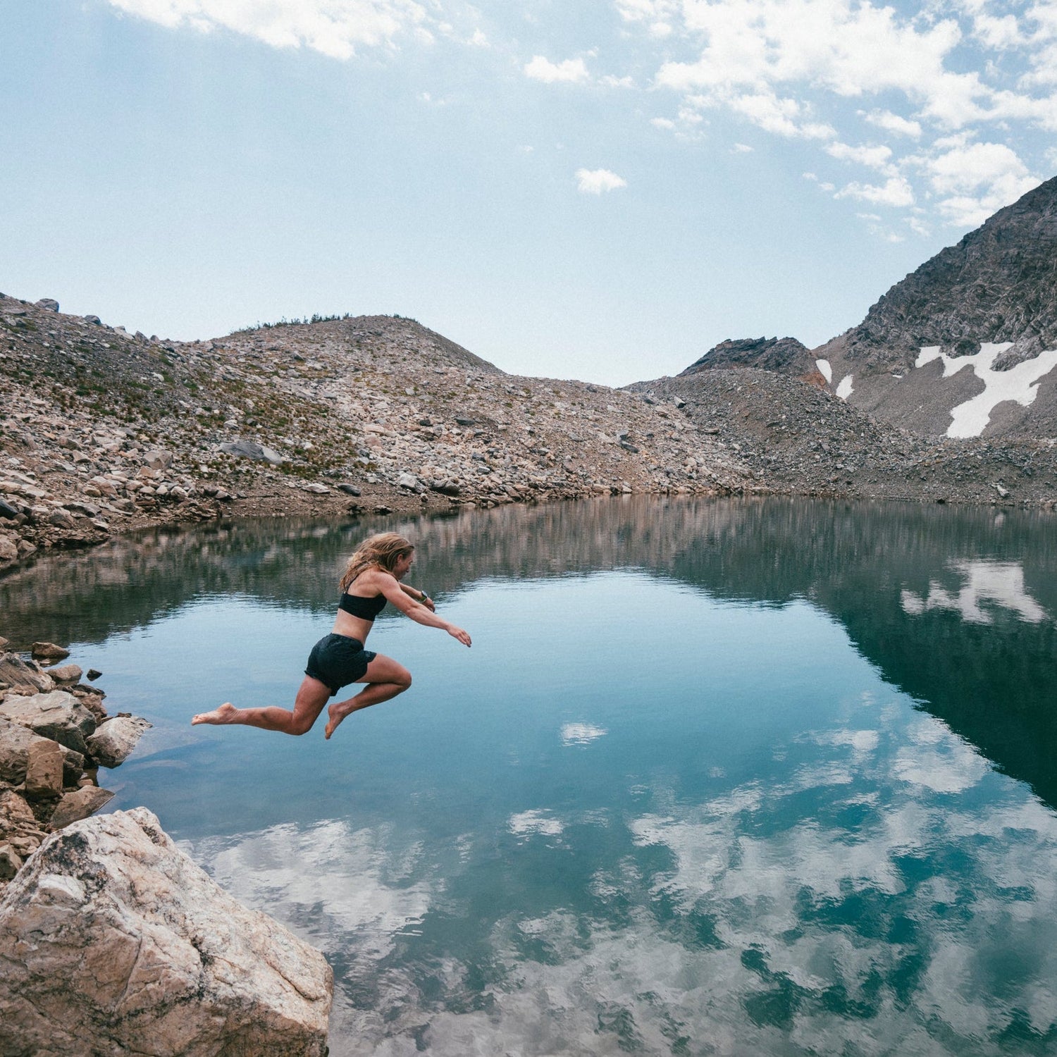 Person jumping into a mountain lake with rocky terrain and snow-capped mountains in the background.