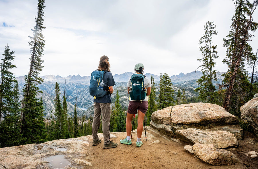 Two hikers standing on a rocky mountain trail overlooking a scenic valley, each wearing a hydration backpack. The scene is framed by tall pine trees and distant mountains under a cloudy sky.