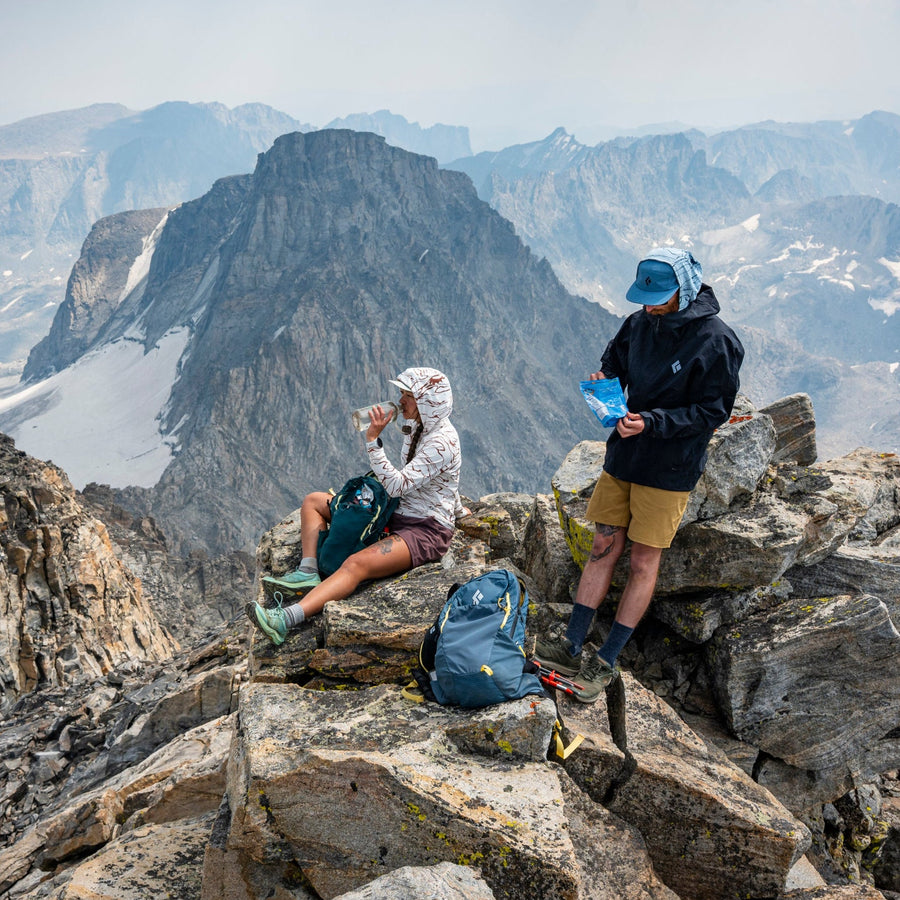 Two hikers on top of a summit having a snack, a mountain vista behind them.