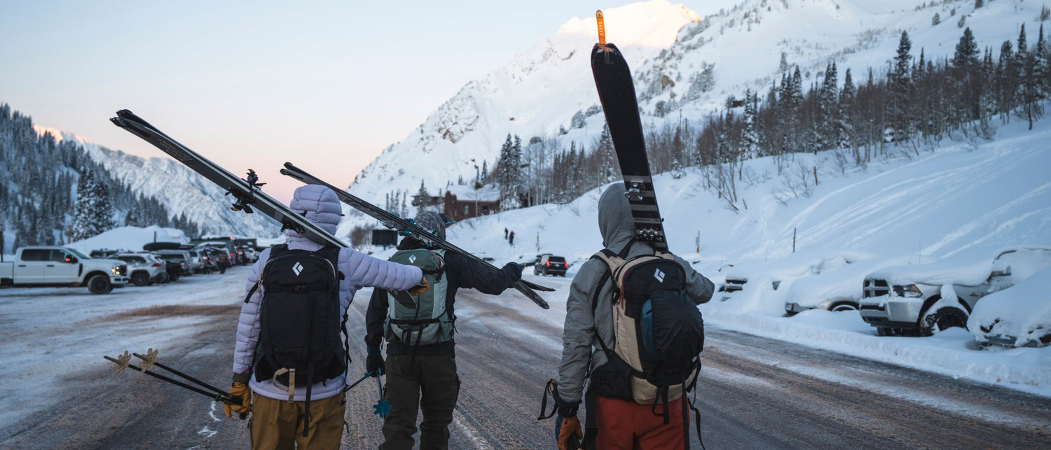 Three people carrying skis on a snowy mountain road with snow-covered trees and mountains in the background.